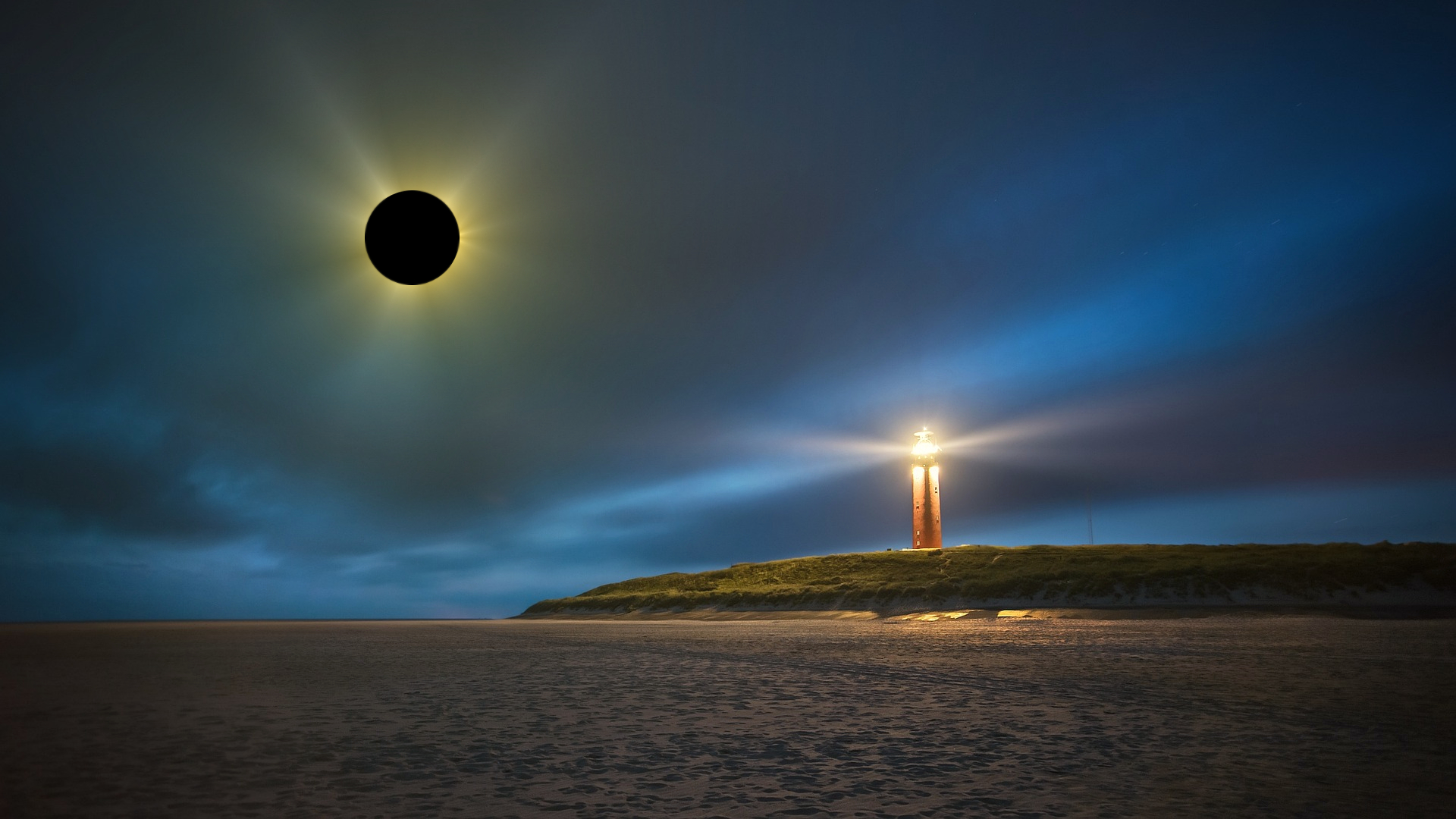 A lighthouse illuminates a seaside beach amid dark skies of subtle blue during the Solar Eclipse.